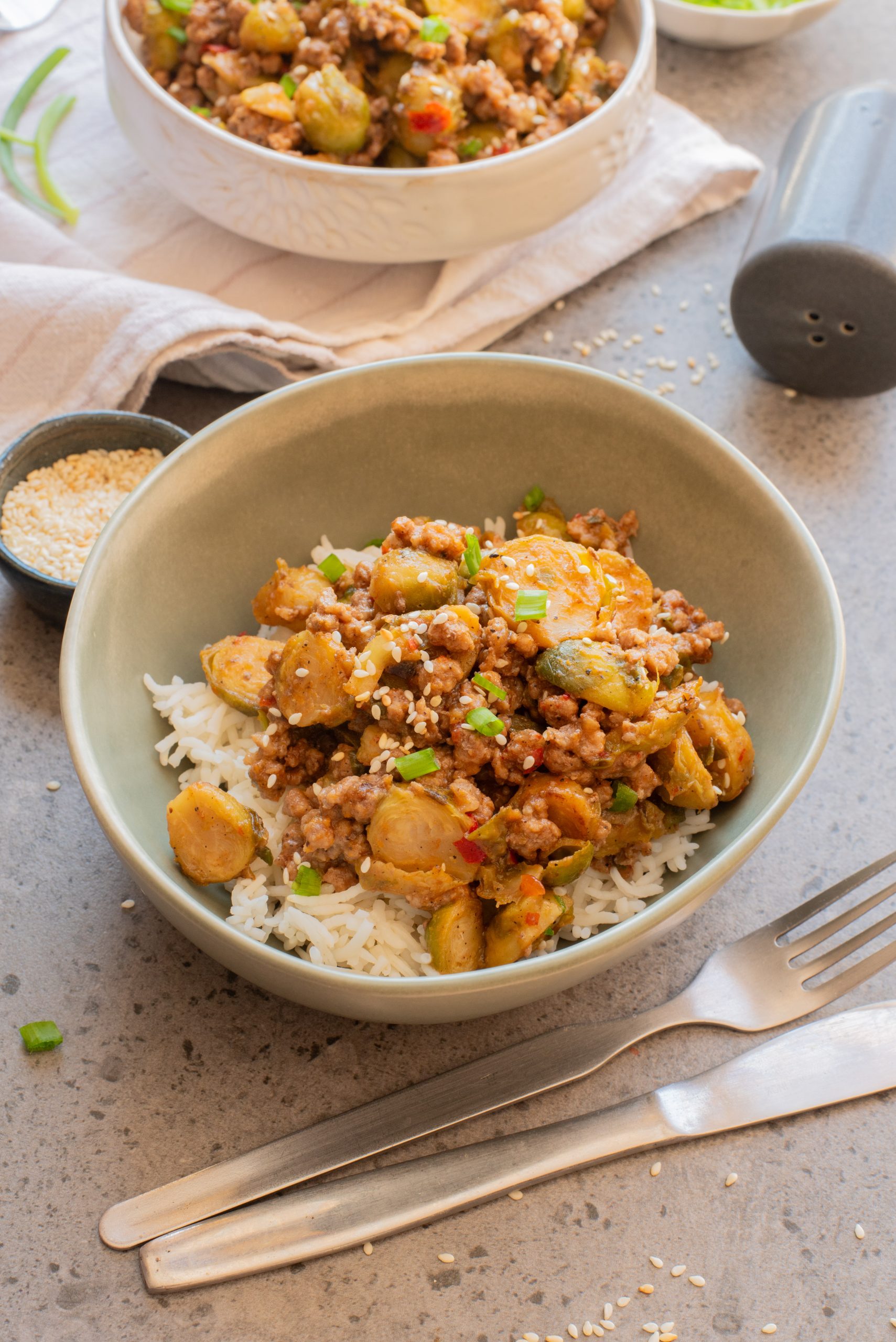 A bowl of white rice topped with a mixture of ground meat, Brussels sprouts, and diced vegetables, garnished with green onions and sesame seeds. A fork and knife are placed beside the bowl.