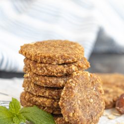 A stack of round, oat-based cookies on parchment paper with a sprig of mint, set against a striped cloth background.