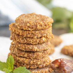 A stack of round, brown cookies sits on parchment paper next to a sprig of mint and a date.