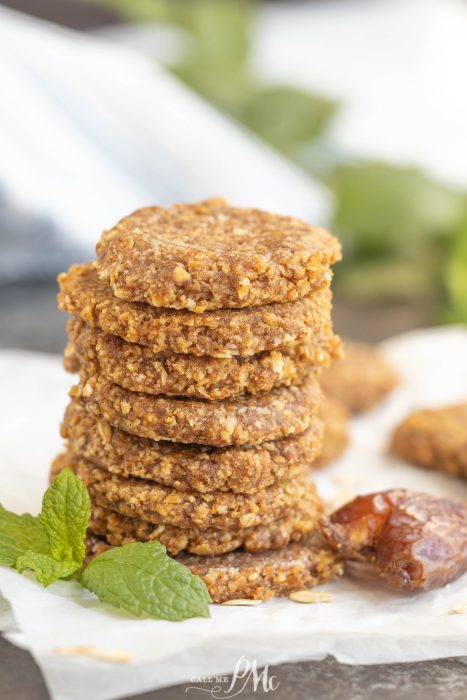 A stack of round, brown cookies sits on parchment paper next to a sprig of mint and a date.