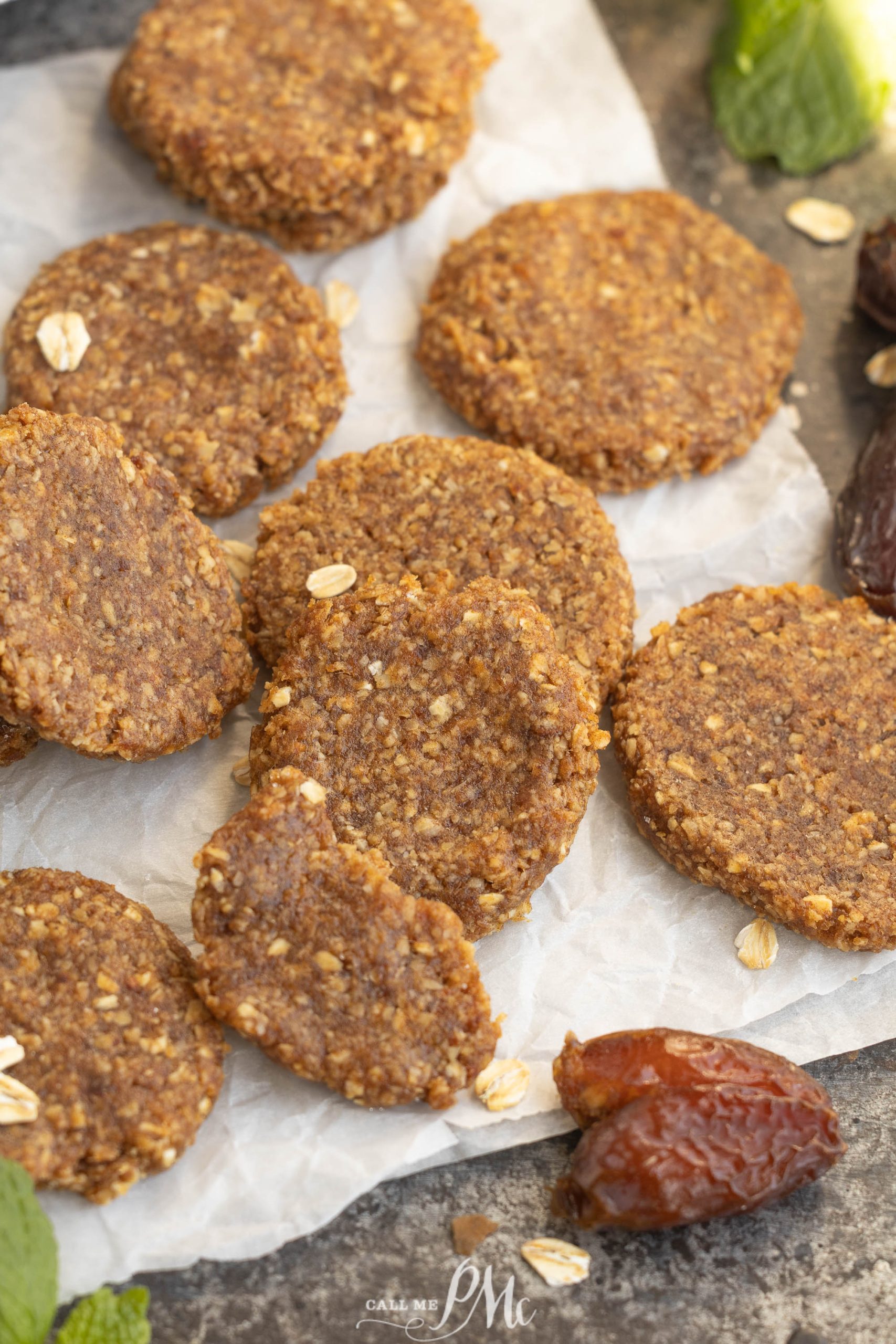 Several round, brown no-bake cookies with a grainy texture are arranged on parchment paper, surrounded by oats and a few dates on a gray surface.