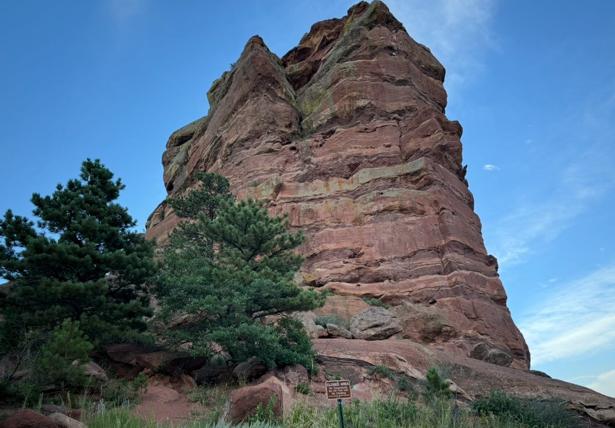 Large red sandstone rock formation stands next to green pine trees under a blue sky; a small brown sign is visible at the base—an iconic stop on some of the best U.S. road trips.