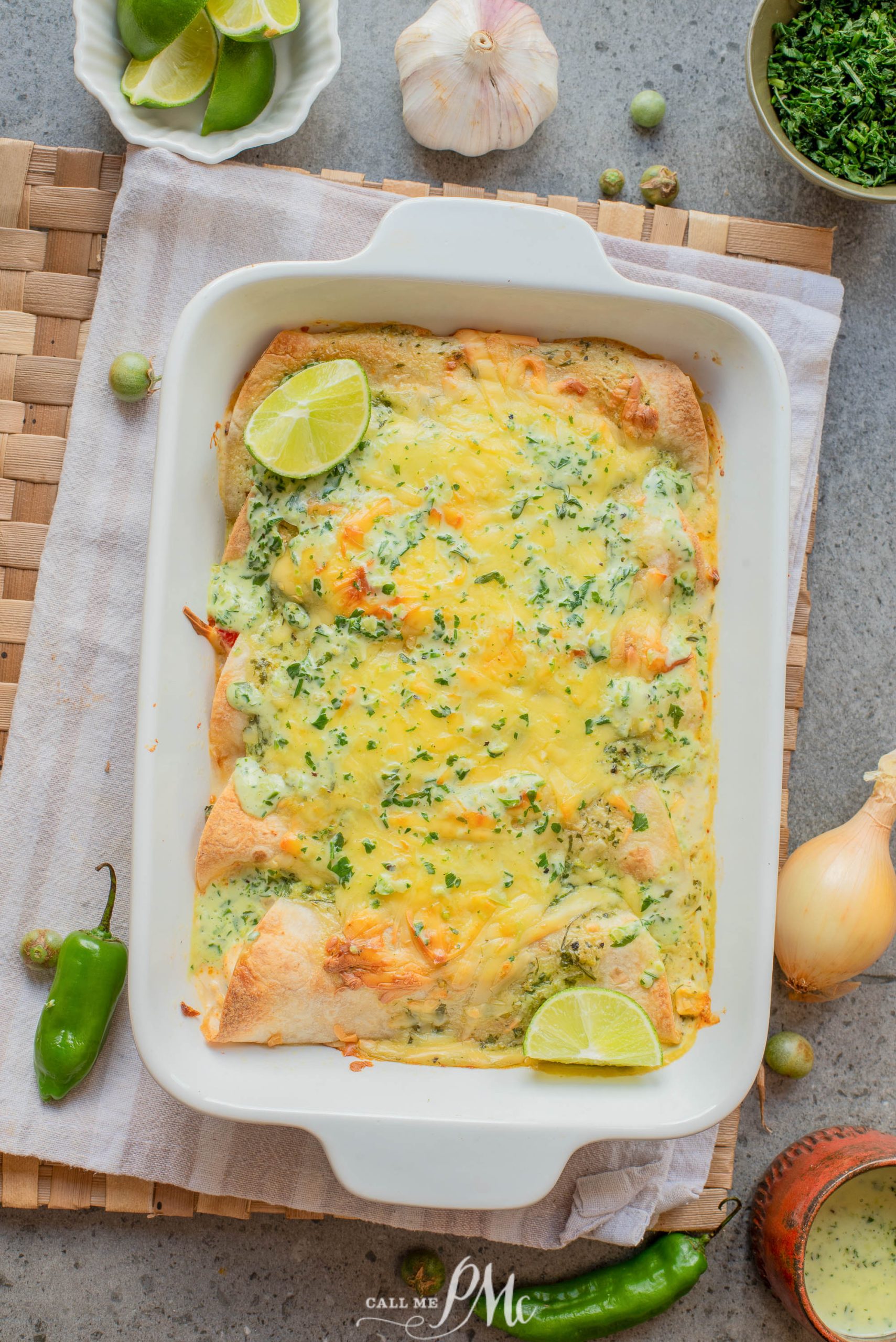 A baking dish filled with cheesy enchiladas topped with chopped herbs and lime wedges, surrounded by fresh ingredients like jalapeรฑo, onion, garlic, and a bowl of limes.