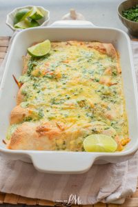 A baked casserole dish filled with cheesy enchiladas, topped with chopped herbs and lime wedges, sits on a cloth napkin.