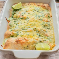 A baked casserole dish filled with cheesy enchiladas, topped with chopped herbs and lime wedges, sits on a cloth napkin.