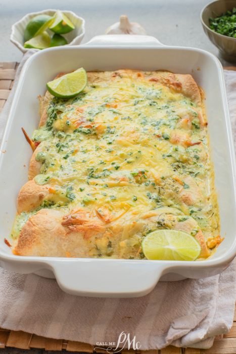 A baked casserole dish filled with cheesy enchiladas, topped with chopped herbs and lime wedges, sits on a cloth napkin.
