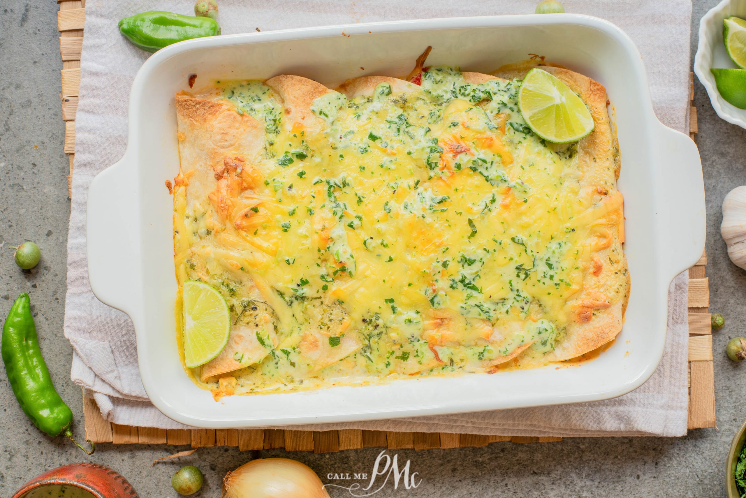 A baked Low-carb shrimp enchiladas casserole dish with melted cheese, green herbs, and lime wedges on top, sitting on a wooden board with onions, peppers, and garlic nearby.