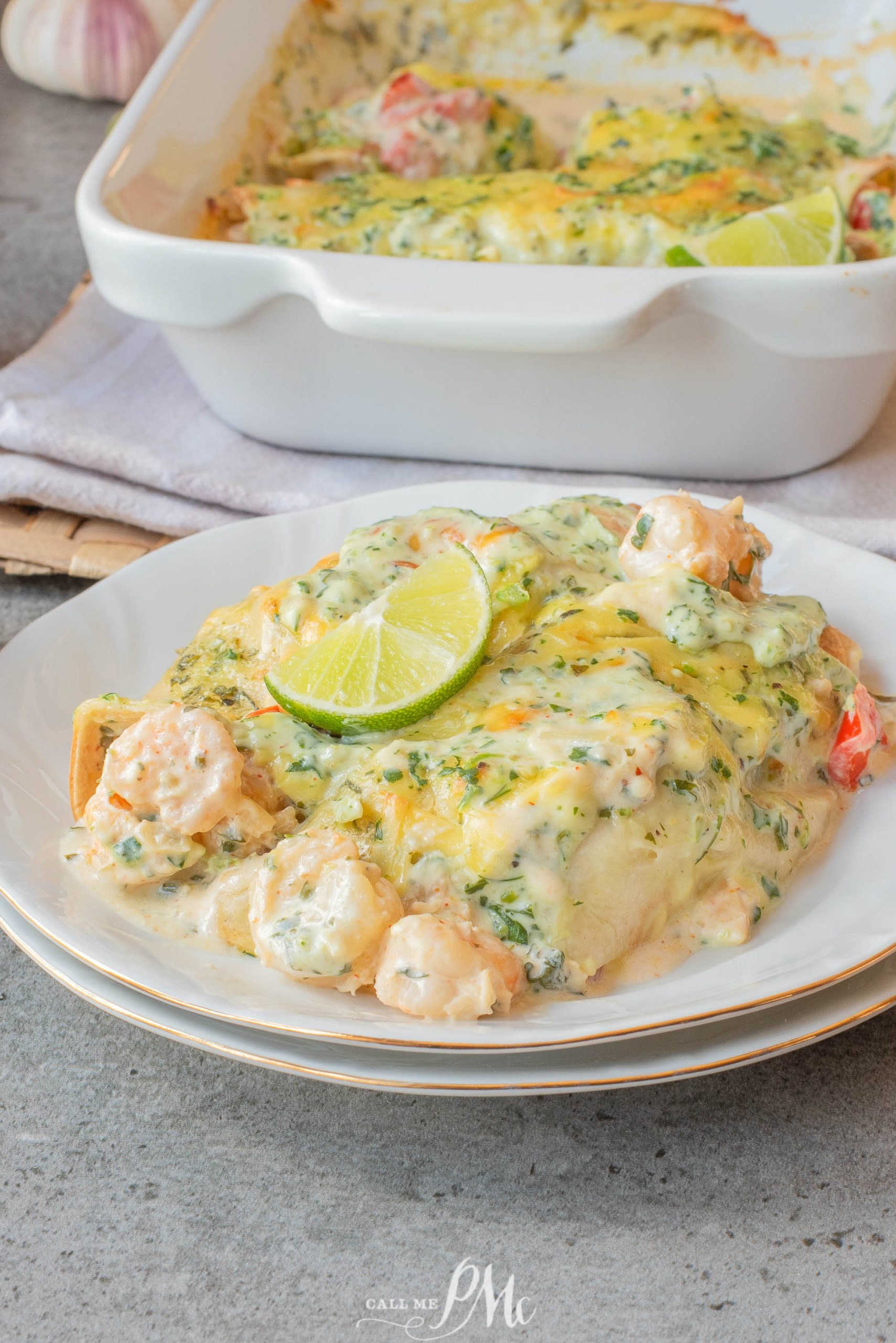 A serving of creamy shrimp casserole topped with melted cheese and a lime slice on a white plate, with a baking dish of casserole in the background.