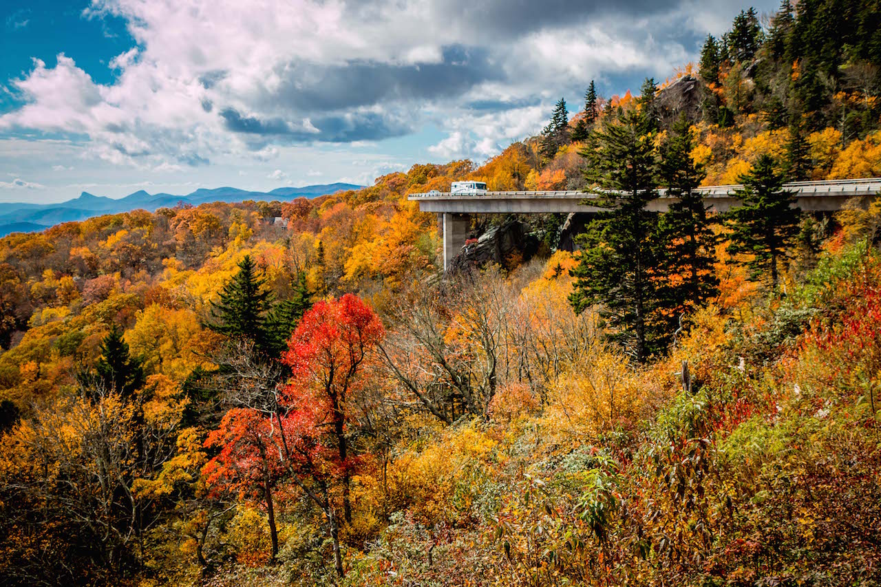 A truck drives over an elevated highway surrounded by colorful autumn trees and distant mountains under a partly cloudy sky, capturing the spirit of the best U.S. road trips.