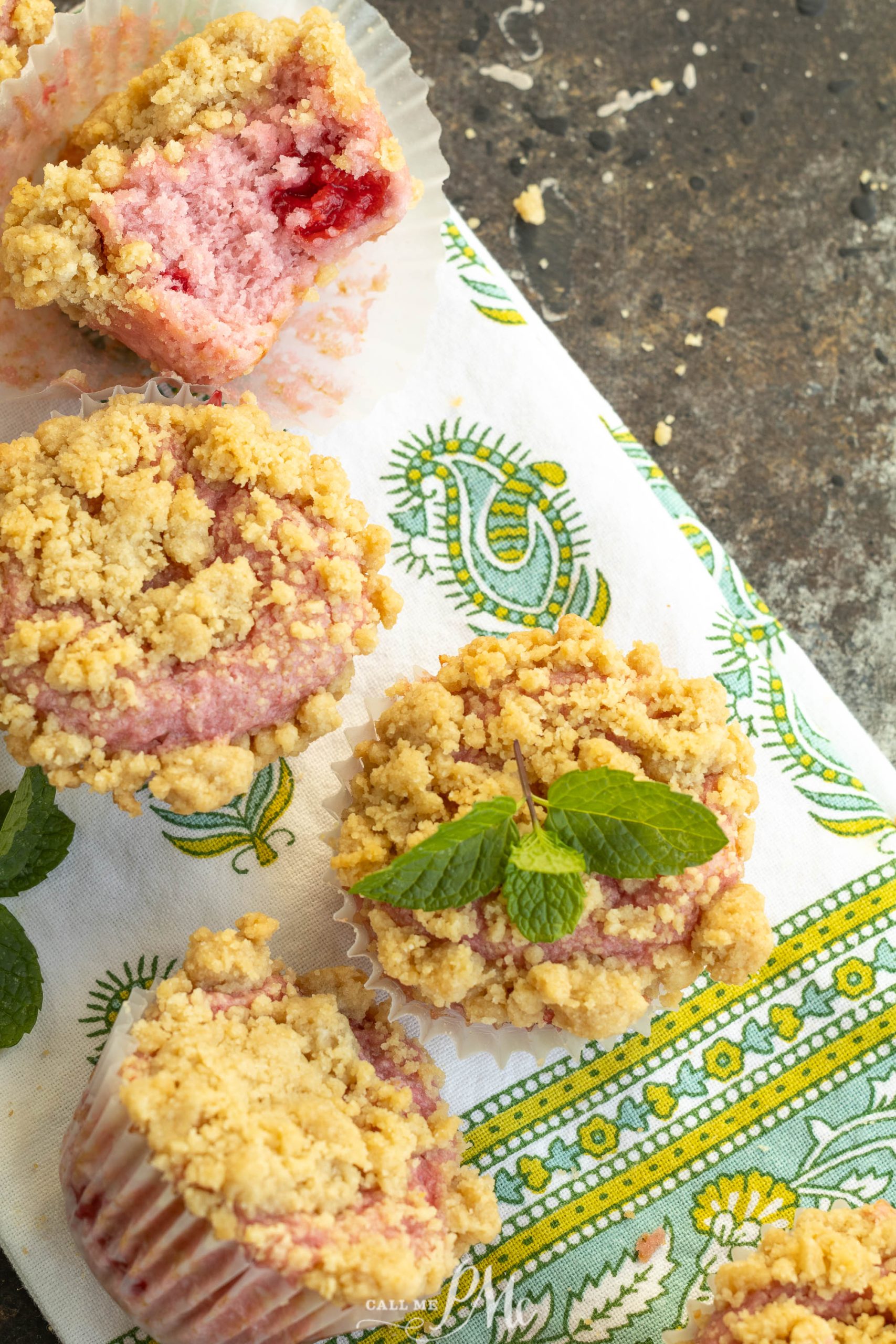 Four crumb-topped cherry muffins, one with a bite taken out, sit on a patterned cloth with a sprig of fresh mint.