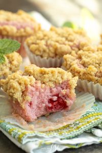 A close-up of crumb-topped pink muffins with a raspberry center, one partially eaten, on a patterned cloth with a mint garnish in the background.