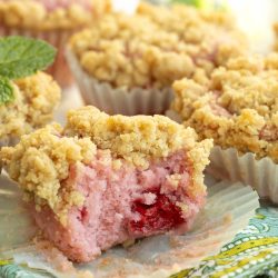 A close-up of crumb-topped pink muffins with a raspberry center, one partially eaten, on a patterned cloth with a mint garnish in the background.