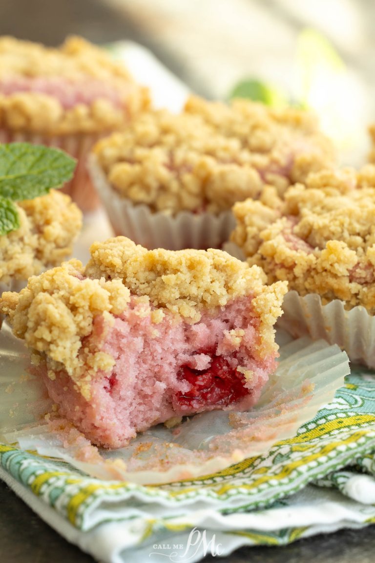 A close-up of crumb-topped pink muffins with a raspberry center, one partially eaten, on a patterned cloth with a mint garnish in the background.