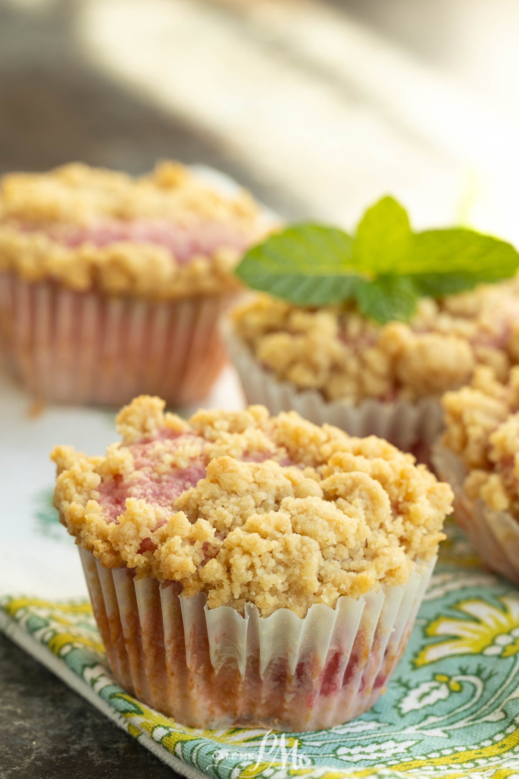 Four crumb-topped muffins, one garnished with a sprig of mint, are arranged on a patterned cloth.