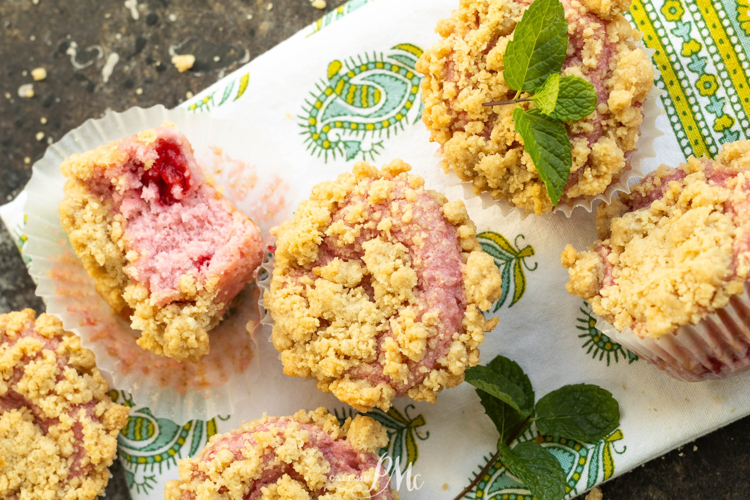 Five strawberry crumble muffins, one with a bite taken out, rest on a patterned cloth; a sprig of mint garnishes one muffin and lies nearby.