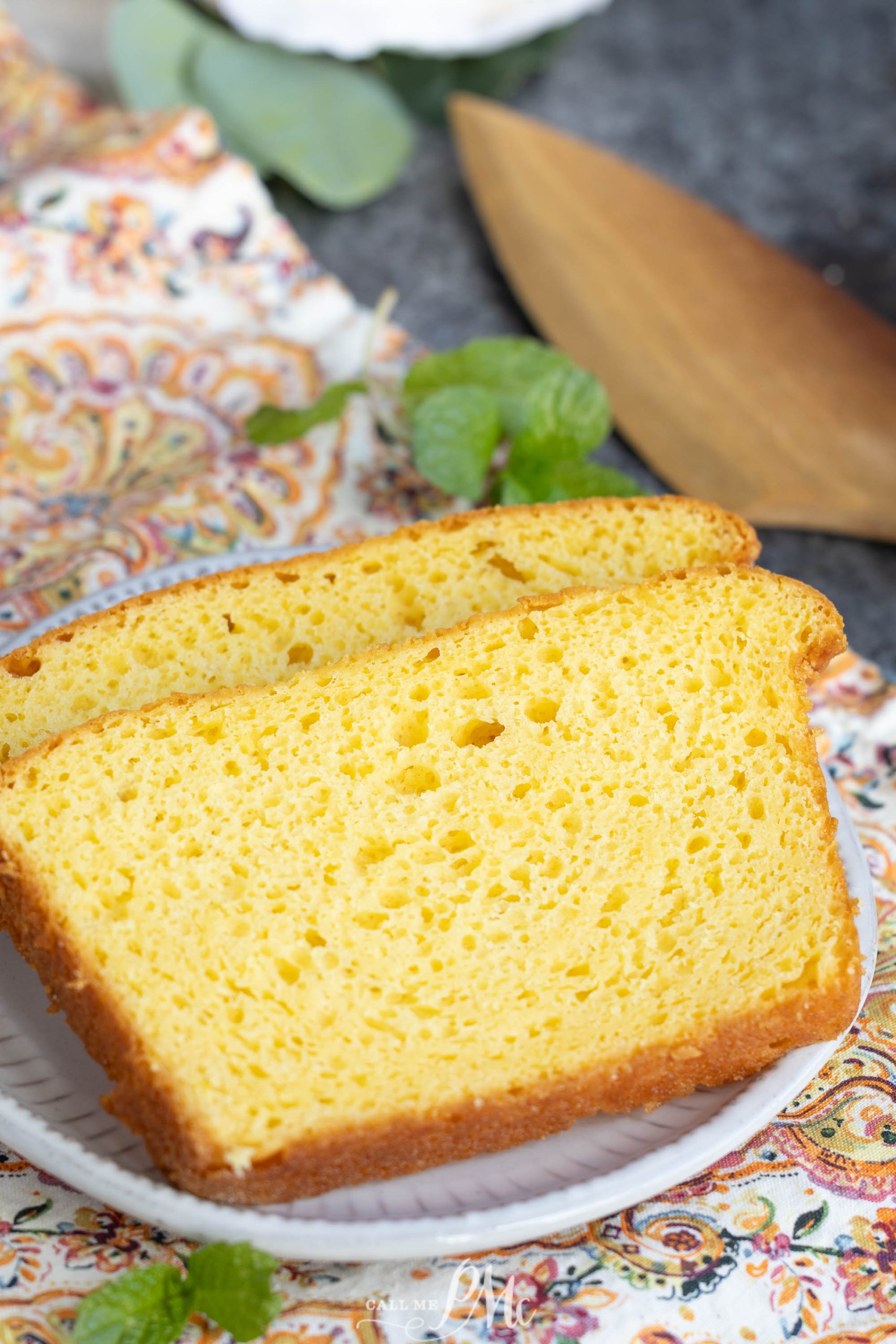Two slices of yellow, airy pound cake are placed on a white plate, set atop a patterned cloth with a wooden spatula and mint leaves nearby.