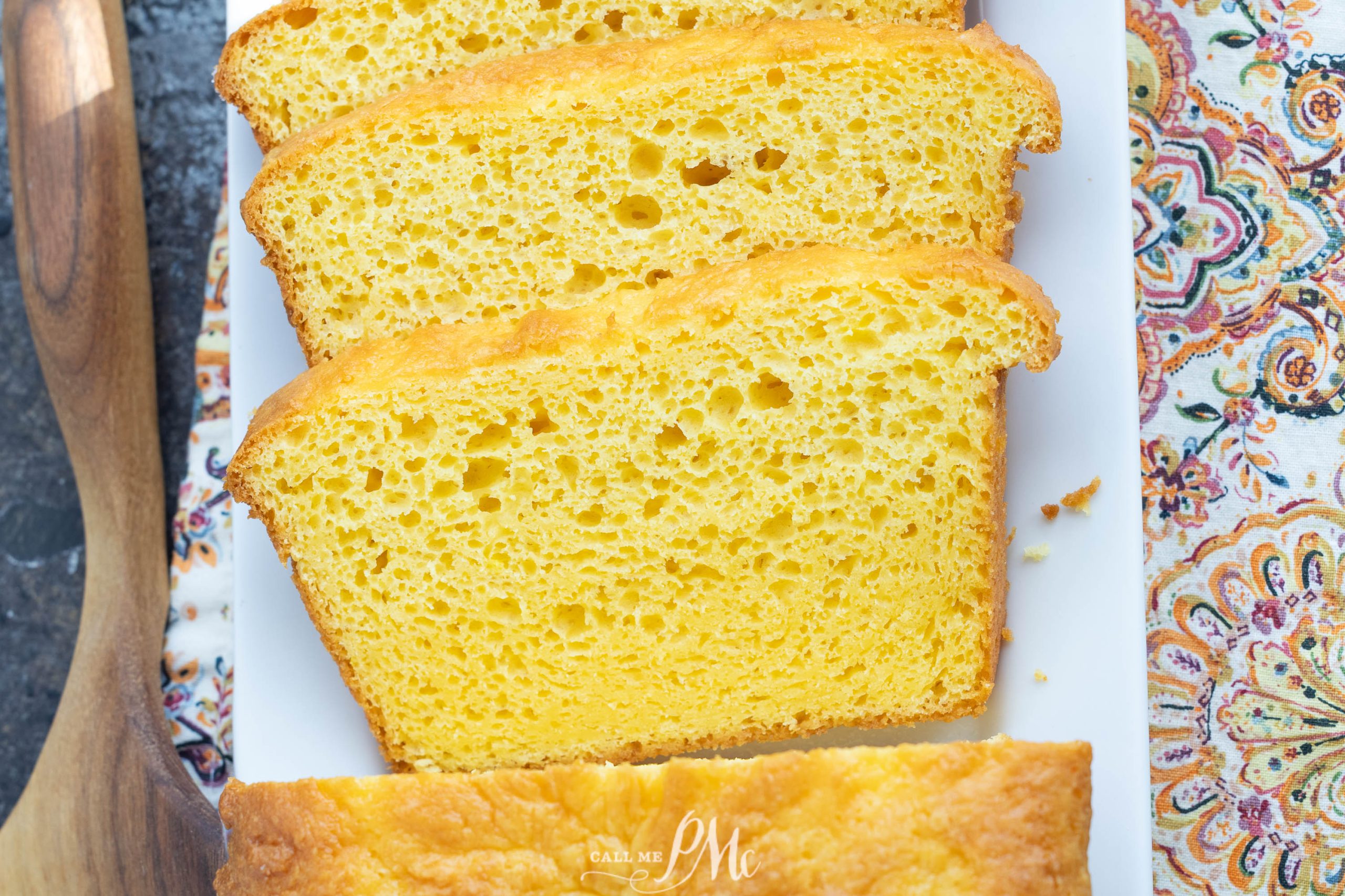 Four slices of yellow cornbread are arranged on a white rectangular plate, next to a wooden spoon and a patterned fabric napkin.
