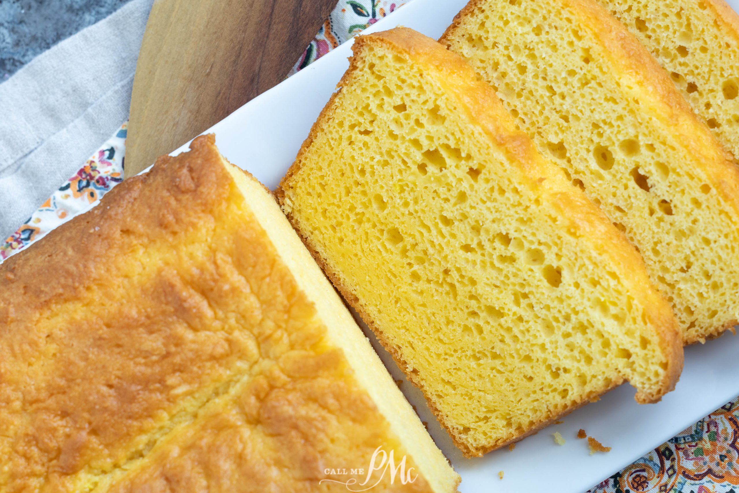 Loaf of cornbread partially sliced on a white rectangular plate, showing its moist, yellow interior and golden crust.