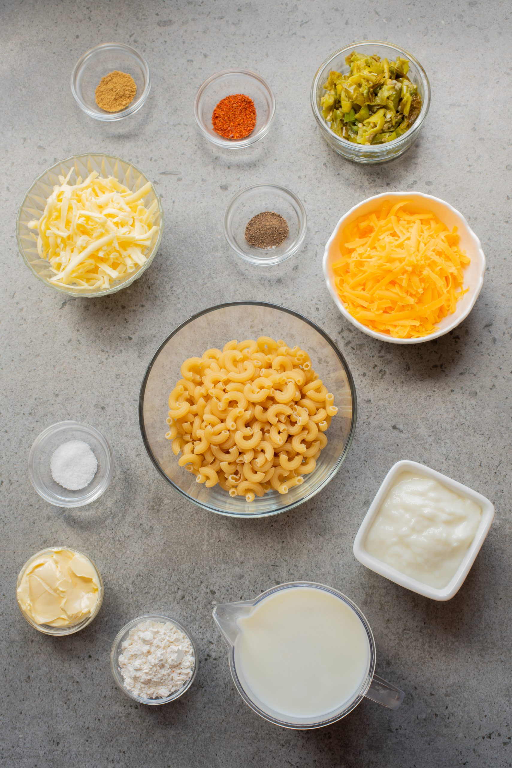 Bowls and containers with ingredients for macaroni and cheese, including elbow pasta, shredded cheese, milk, butter, yogurt, flour, spices, pickled jalapeños, and salt on a gray surface.