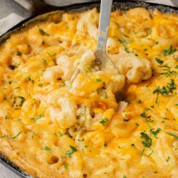 A skillet filled with baked macaroni and cheese topped with melted cheddar, being served with a metal spoon and garnished with chopped parsley.