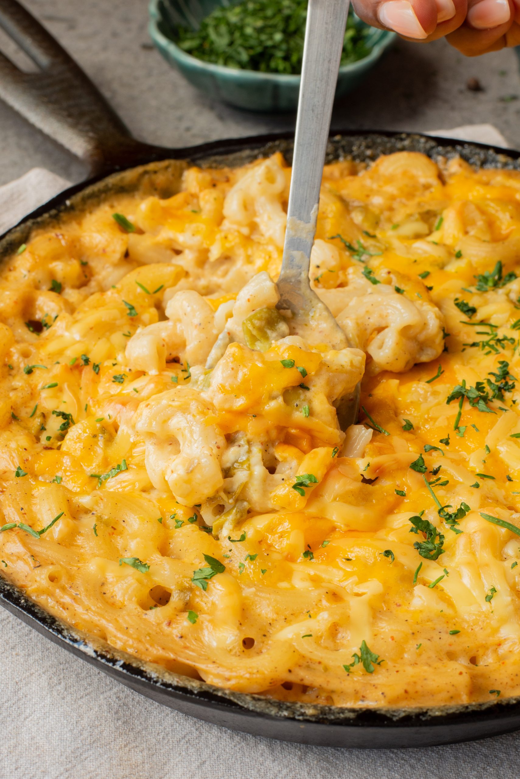 A skillet filled with baked macaroni and cheese topped with melted cheddar, being served with a metal spoon and garnished with chopped parsley.