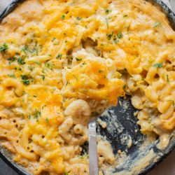 A skillet filled with baked macaroni and cheese, partially served, topped with melted cheddar and garnished with chopped herbs.