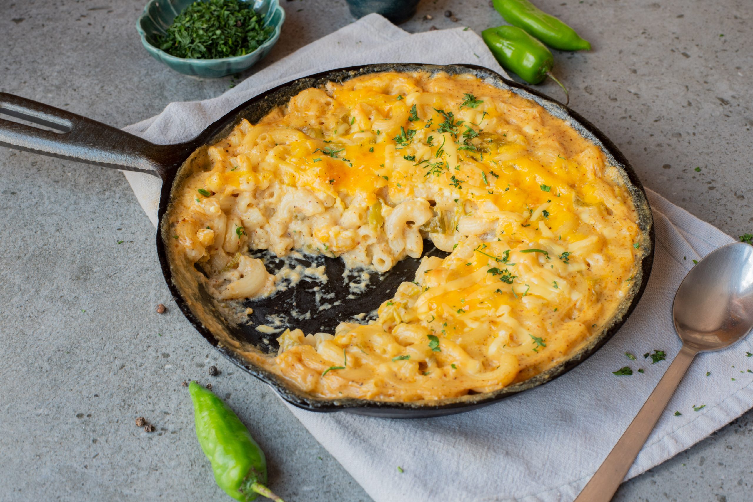 A cast iron skillet with baked macaroni and cheese, partially served, garnished with herbs, on a cloth napkin with a spoon and green chili peppers nearby.
