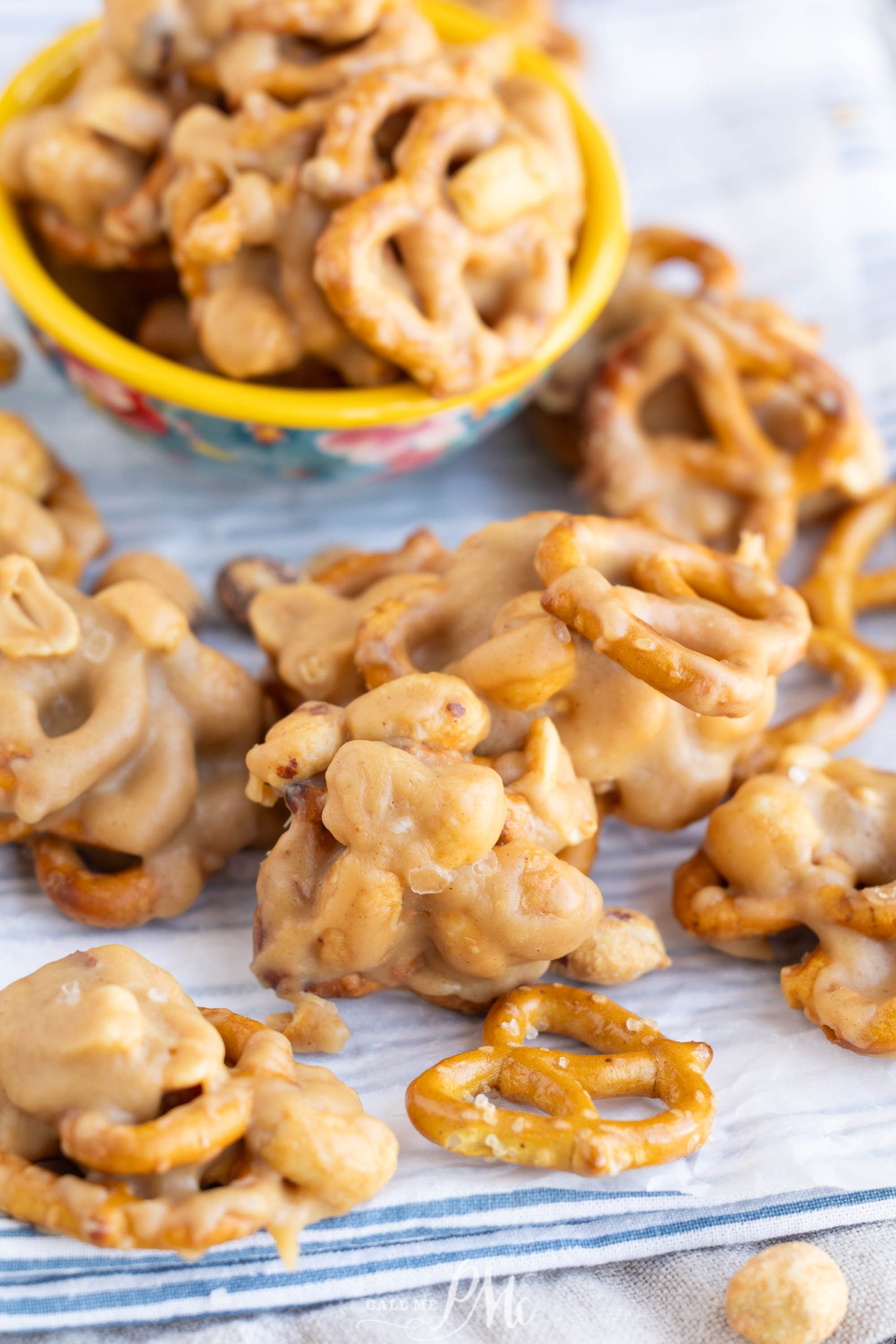 Clusters of pretzels and peanuts coated in a light brown candy or caramel coating are arranged on a striped cloth, with a yellow bowl holding more clusters in the background.