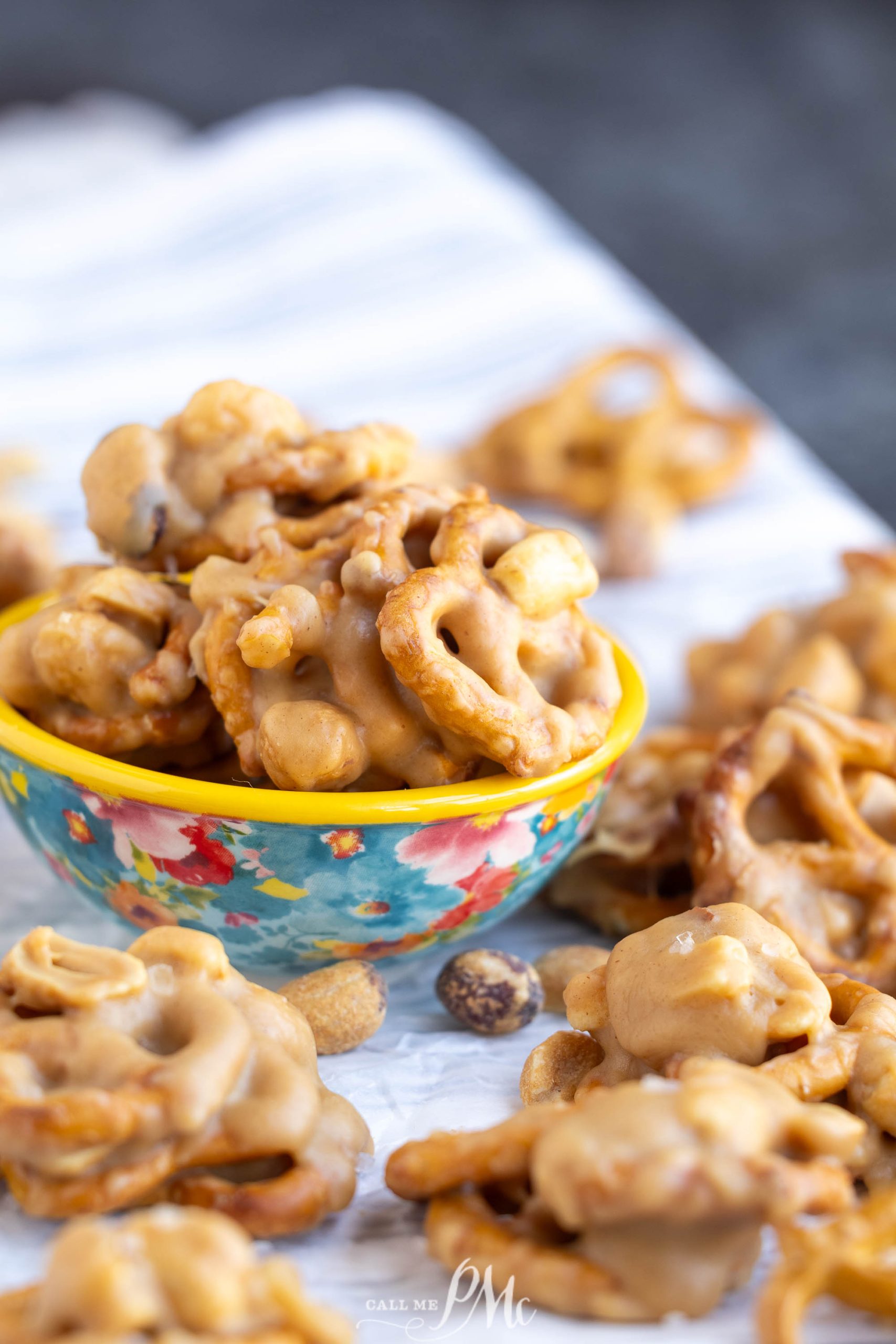 A small floral bowl filled with clusters of pretzels and peanuts coated in a light brown candy mixture, with more clusters scattered around on a white surface.