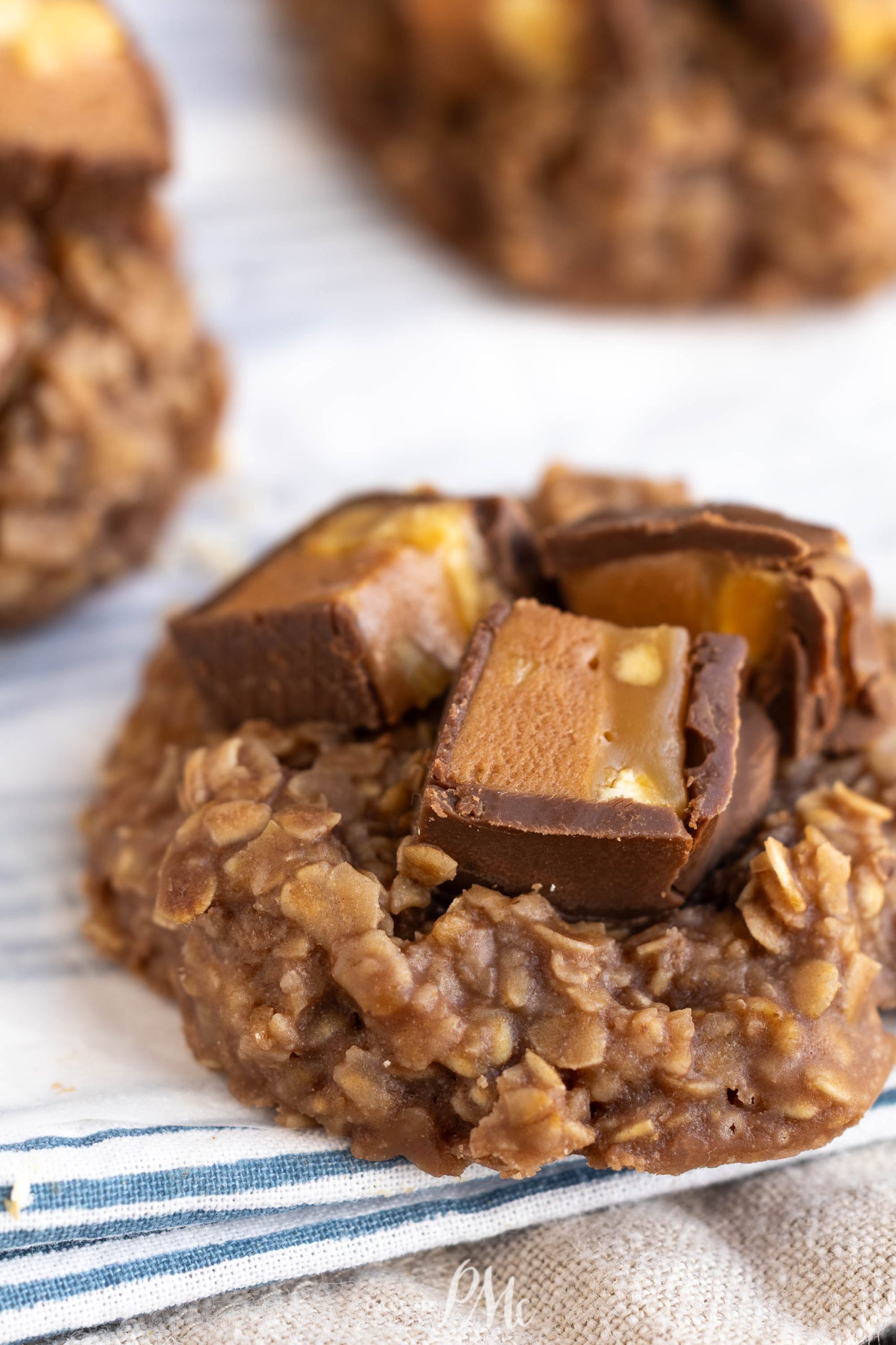 A no-bake chocolate oatmeal cookie topped with chopped pieces of chocolate and caramel candy on a striped cloth.