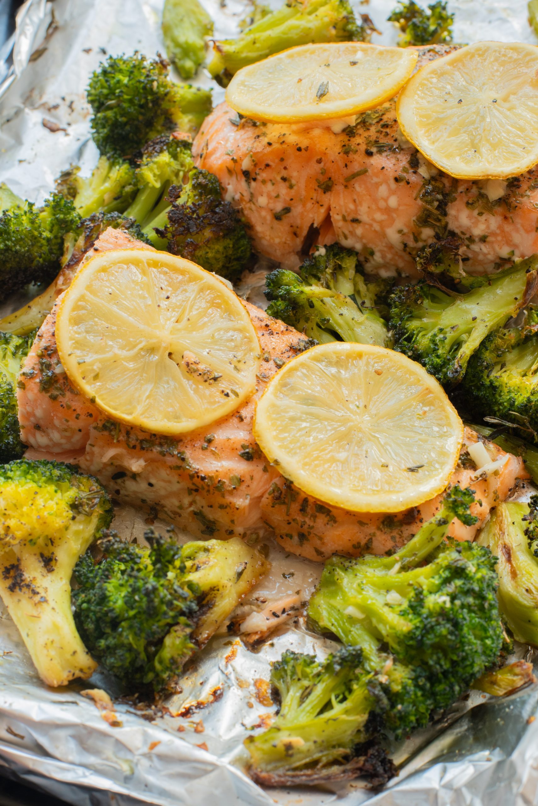 Baked salmon fillets topped with lemon slices and surrounded by roasted broccoli on a foil-lined baking sheet.