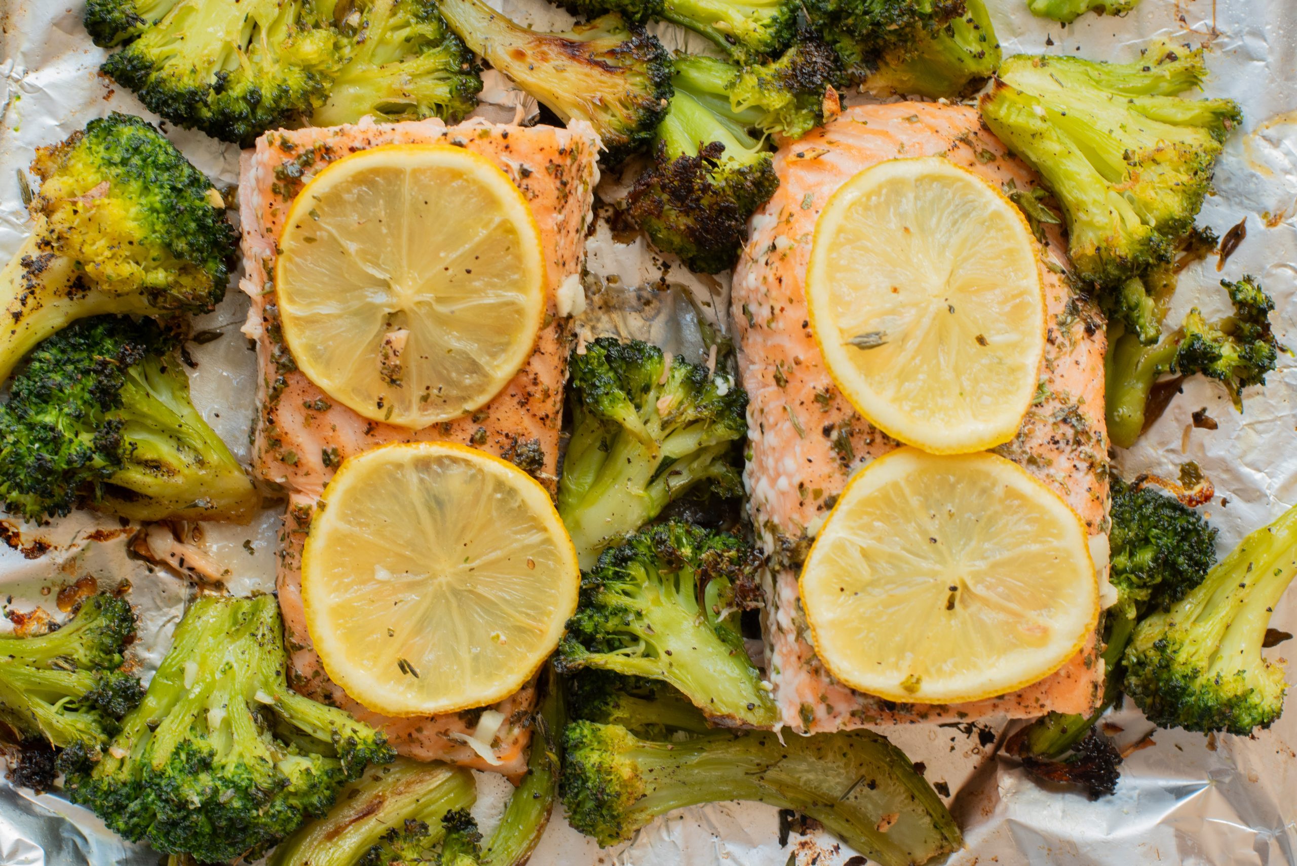 Two baked salmon fillets topped with lemon slices, surrounded by roasted broccoli florets on a sheet of aluminum foil.