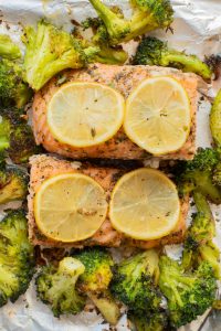 Two baked salmon fillets topped with lemon slices, surrounded by roasted broccoli on a foil-lined baking sheet.