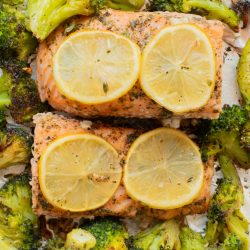 Two baked salmon fillets topped with lemon slices, surrounded by roasted broccoli on a foil-lined baking sheet.