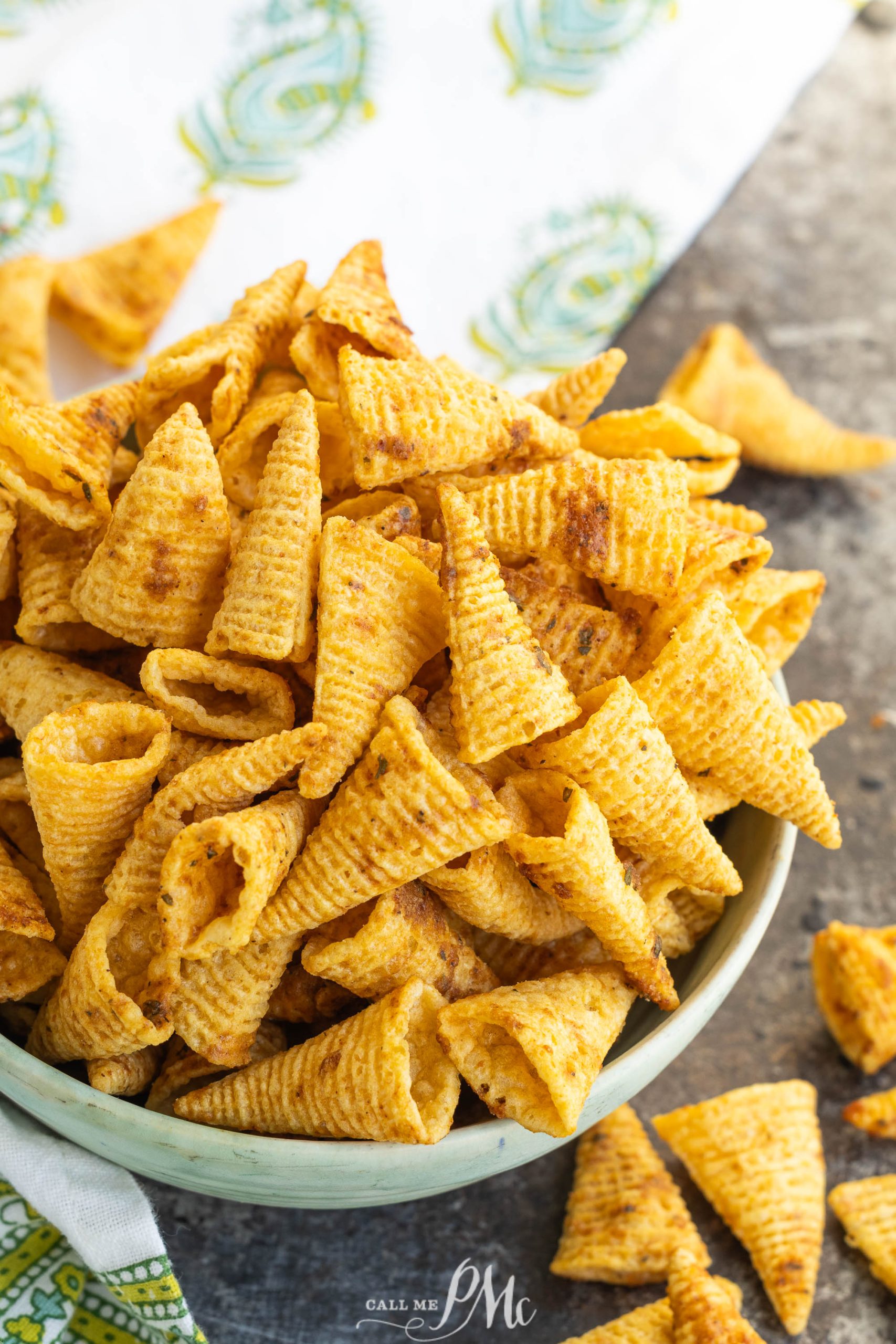 A bowl filled with crunchy, Cheesy Cajun Bugles sits on a textured surface, with a few cone-shaped chips scattered around the bowl.