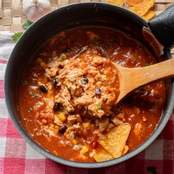 A pot of Chicken Enchilada Soup with beans, corn, shredded meat, and tortilla chips sits on a red and white checkered cloth, with a wooden spoon resting inside.