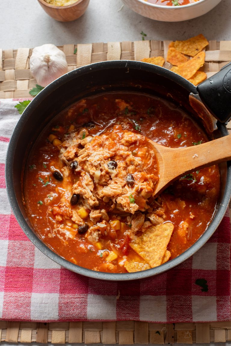 A pot of Chicken Enchilada Soup with beans, corn, shredded meat, and tortilla chips sits on a red and white checkered cloth, with a wooden spoon resting inside.