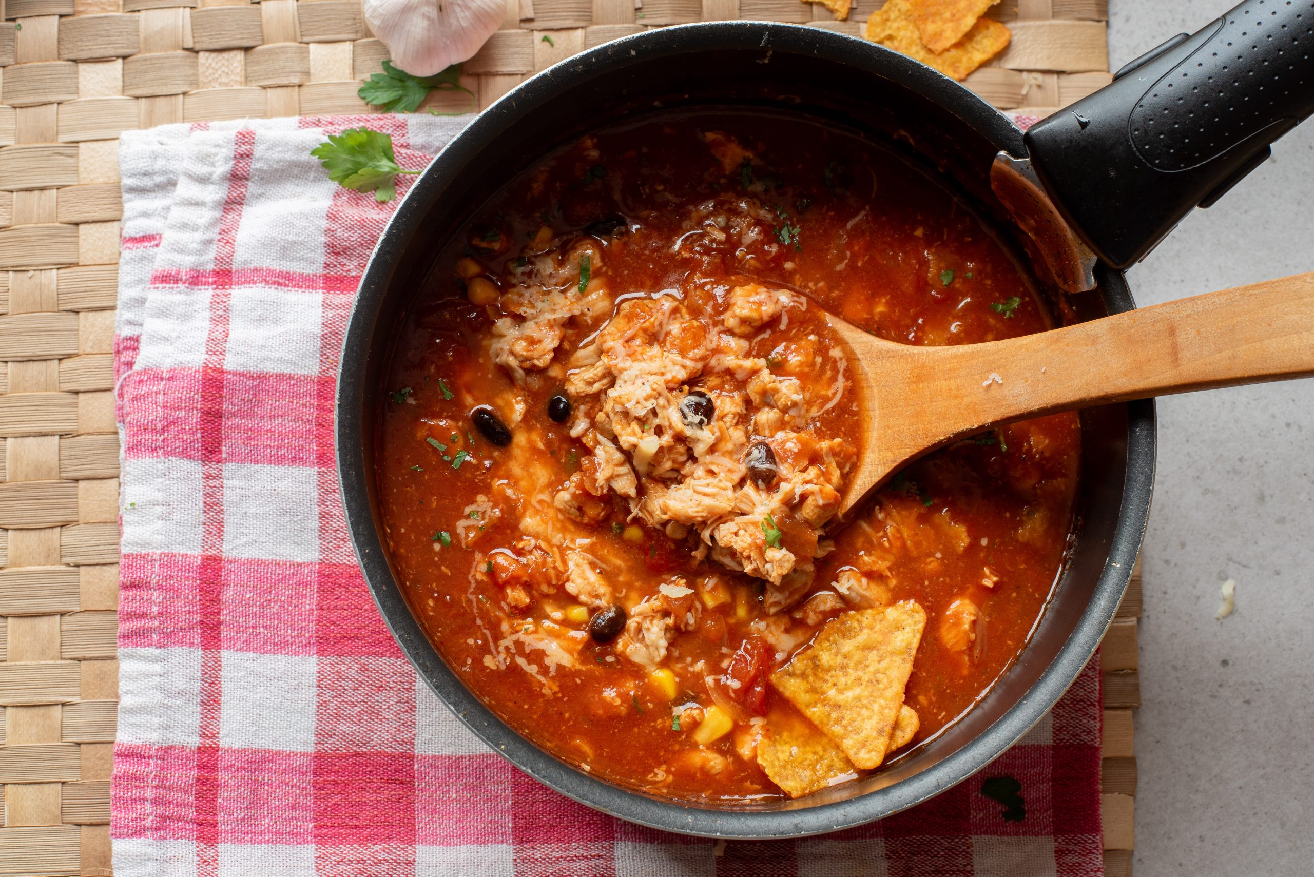 A pot of Chicken Enchilada Soup with black beans and corn, stirred with a wooden spoon, sits on a red and white checkered towel.