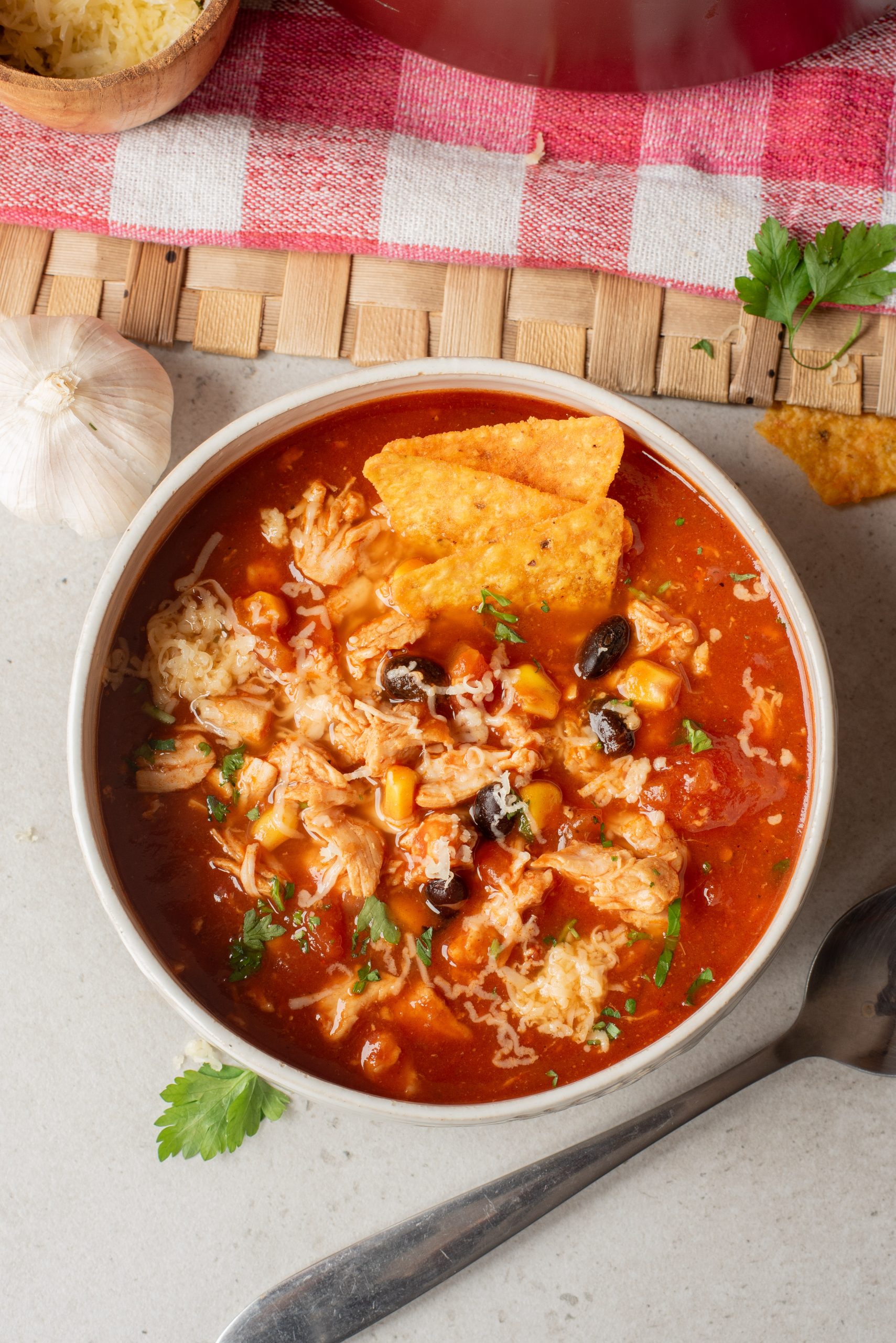 A bowl of Chicken Enchilada Soup topped with shredded cheese, black beans, corn, and tortilla chips sits on a light surface beside a spoon, garlic, and parsley.