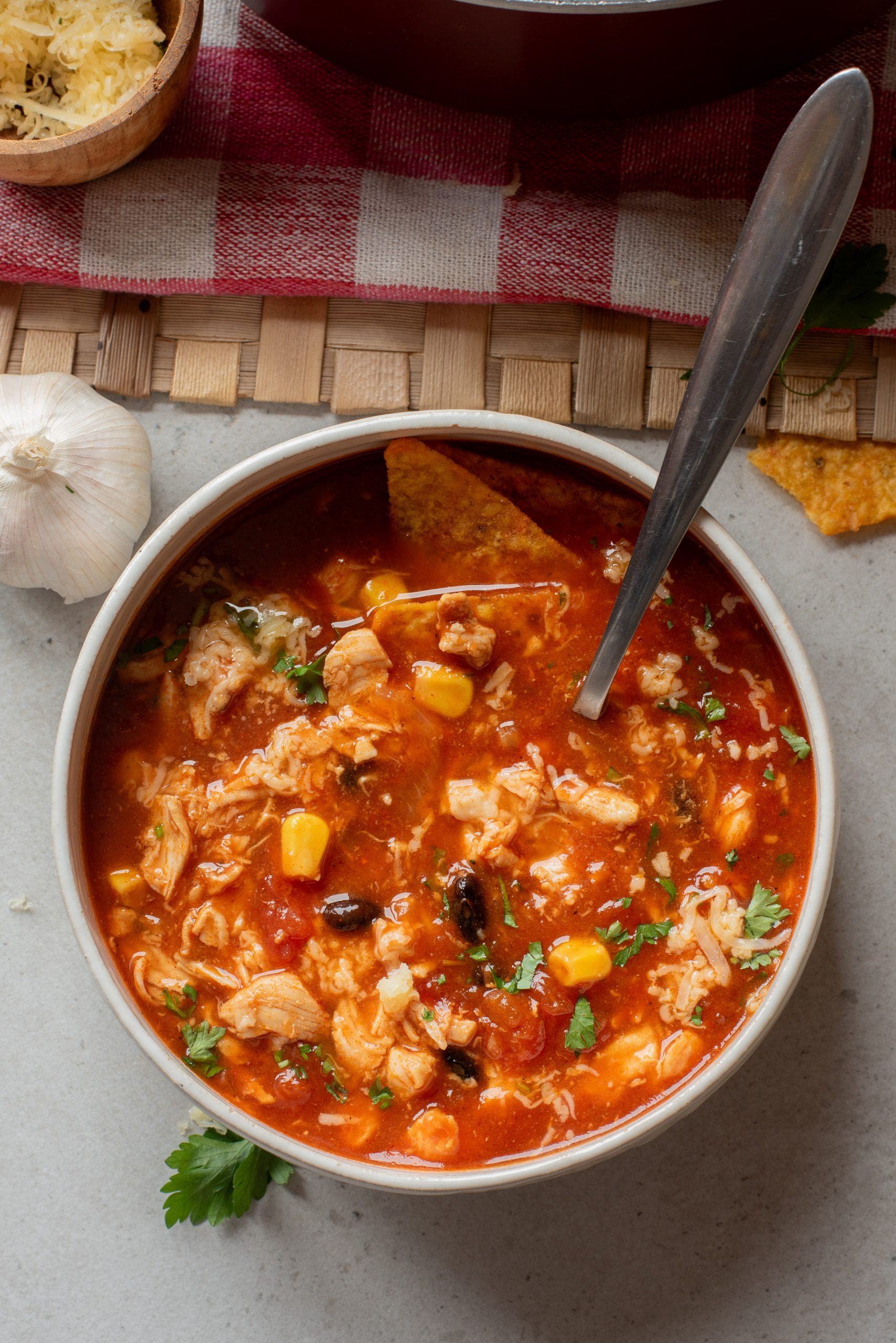 A bowl of Chicken Enchilada Soup with corn, beans, shredded chicken, and a tortilla chip, garnished with herbs, sits on a light surface next to a garlic bulb.
