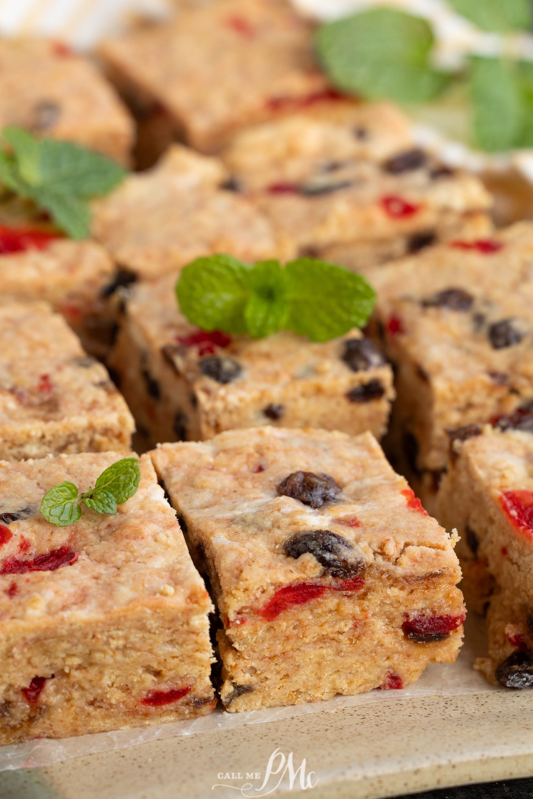 Close-up of several square oat bars with visible red and dark dried fruits, reminiscent of Christmas Icebox Fruitcake, topped with small sprigs of fresh mint and arranged on a plate.