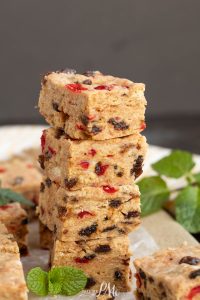 A stack of four beige dessert bars with red and dark brown pieces, reminiscent of Christmas Icebox Fruitcake, sits on parchment paper with mint leaves nearby.
