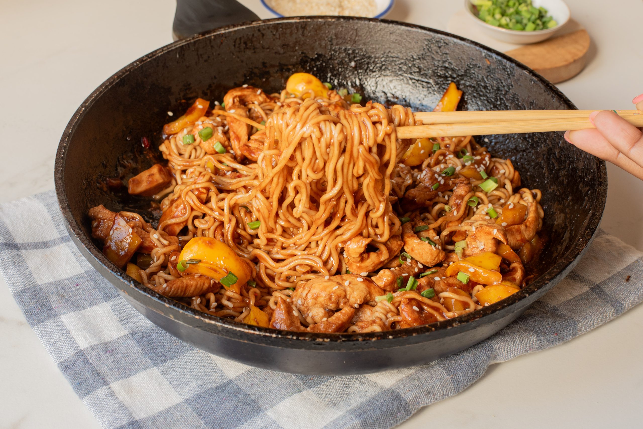 A skillet of Chicken Dragon Noodles stir-fried with chicken, bell peppers, and green onions, with chopsticks lifting a portion, placed on a checkered cloth.