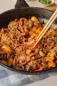 A skillet filled with Chicken Dragon Noodles, sliced chicken, yellow bell peppers, and chopped green onions is being stirred with wooden chopsticks.