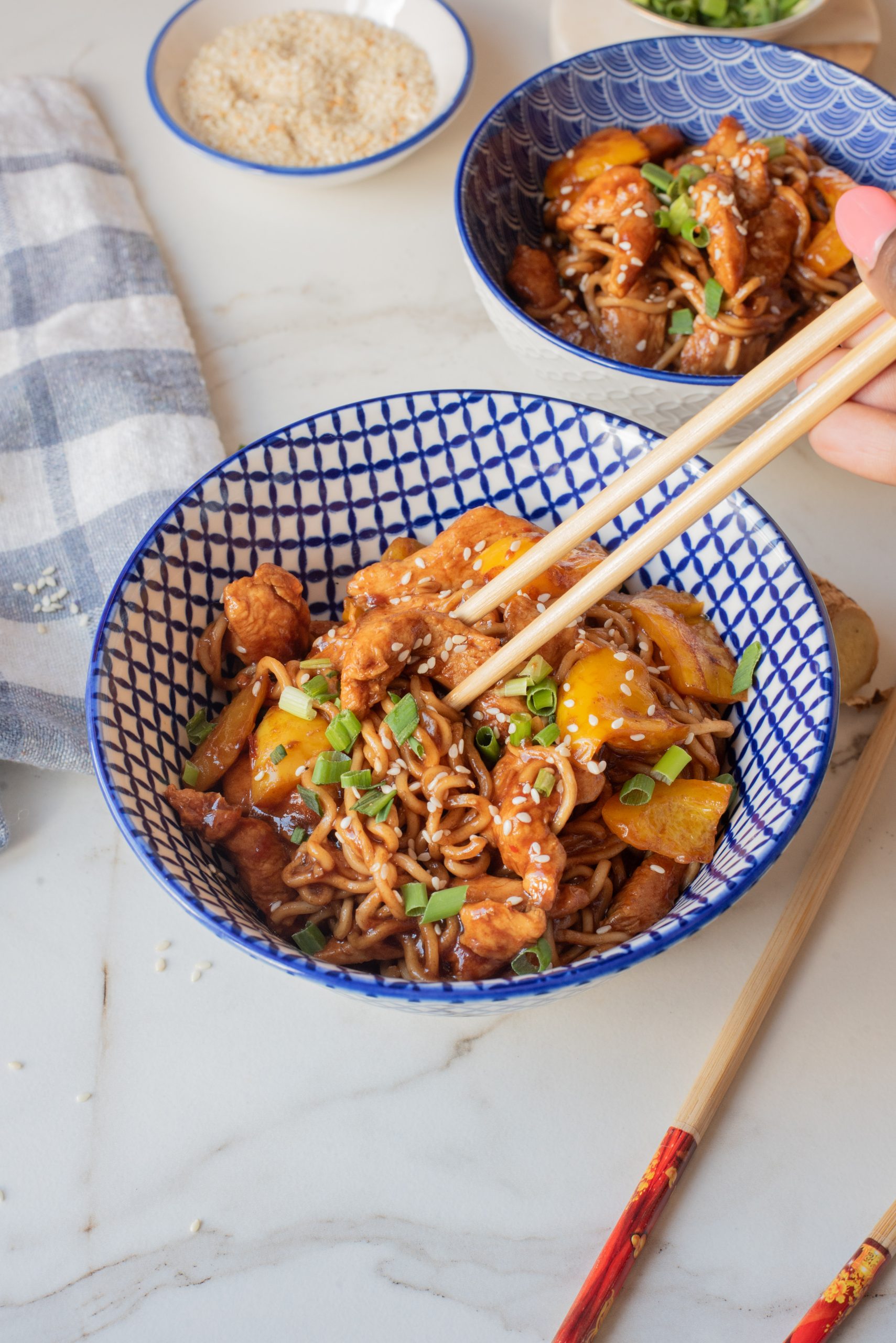 A hand uses chopsticks to pick up Chicken Dragon Noodles with chicken and vegetables from a patterned bowl. Another bowl of the same dish and a small bowl of sesame seeds are also visible.