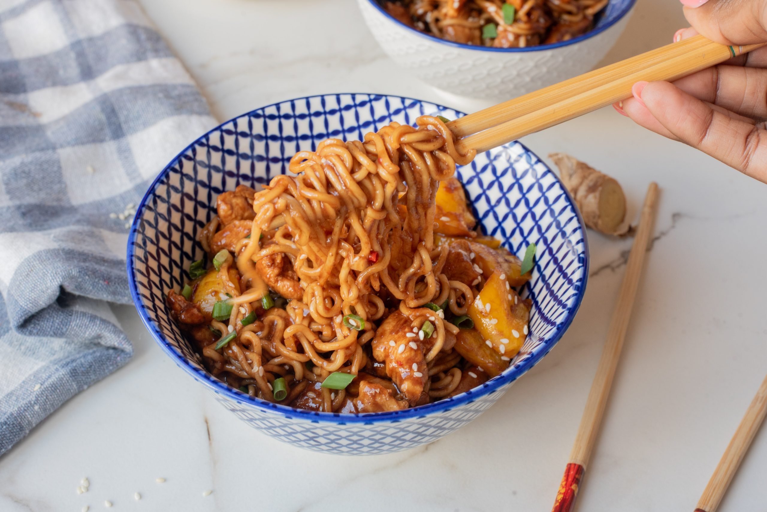 A hand uses chopsticks to lift food from a patterned bowl, set on a marble surface with a napkin and extra chopsticks nearby.