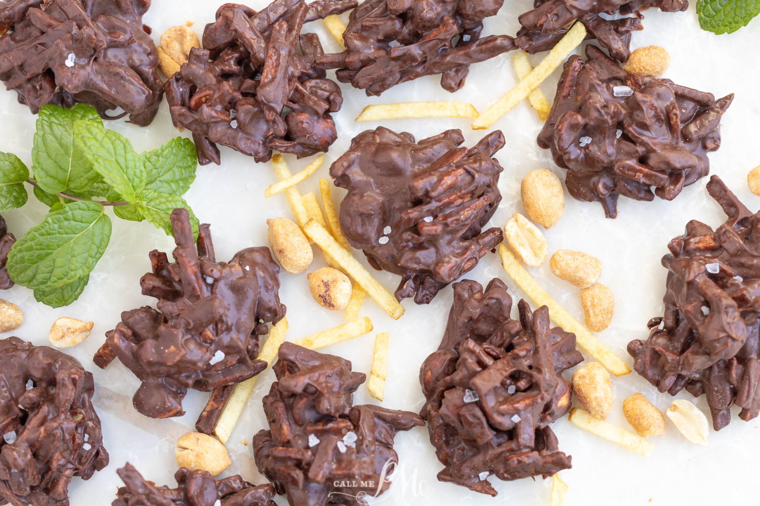 Clusters of chocolate-covered potato sticks, peanuts, and sea salt are arranged on a white surface with scattered peanuts, potato sticks, and mint leaves. Shoestring Potato Chip Haystacks