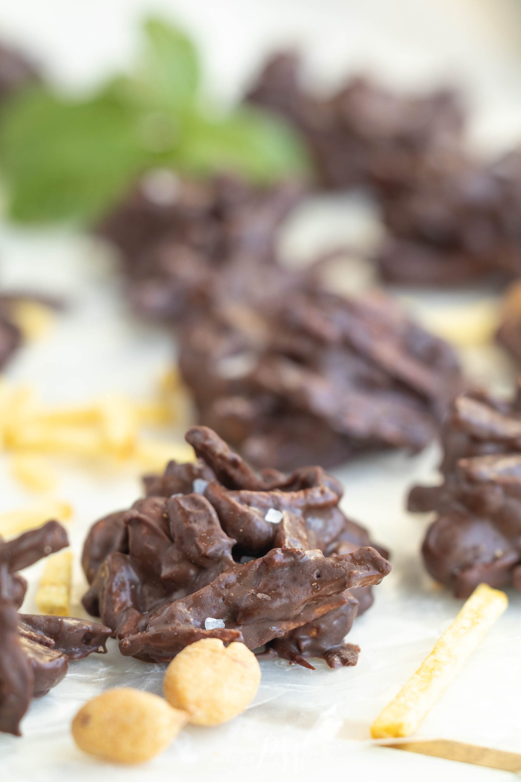 Clusters of chocolate-covered snack sticks and peanuts are arranged on parchment paper, with a few yellow snack sticks and a blurred green garnish in the background.