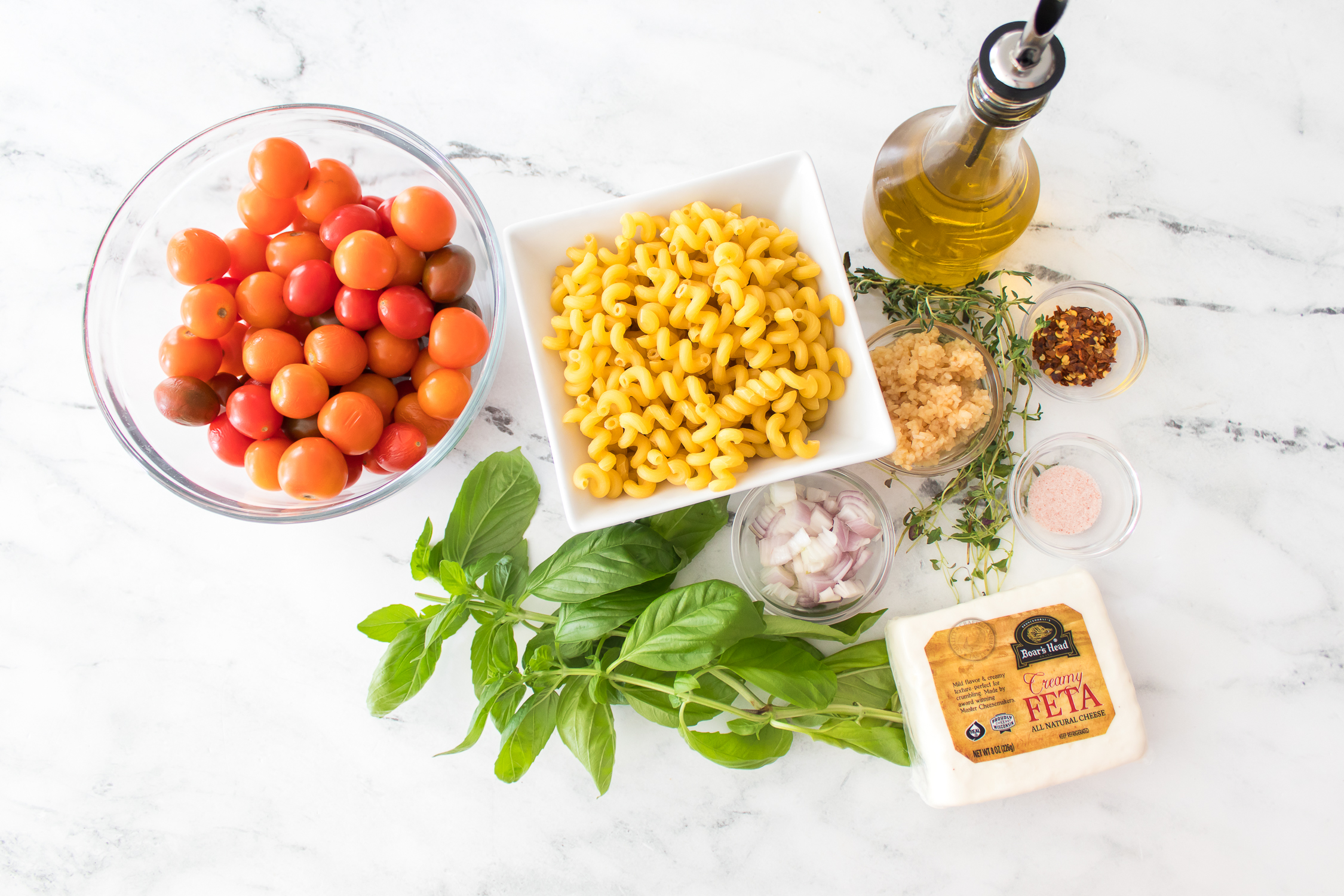 A bowl of cherry tomatoes, a square bowl of cooked pasta, olive oil, feta cheese, fresh basil, shallots, garlic, herbs, red pepper flakes, and salt arranged on a marble surface for Burst Tomato Feta Pasta.