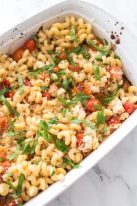 A white baking dish filled with baked pasta, cherry tomatoes, crumbled feta cheese, and fresh basil, set on a marble countertop.